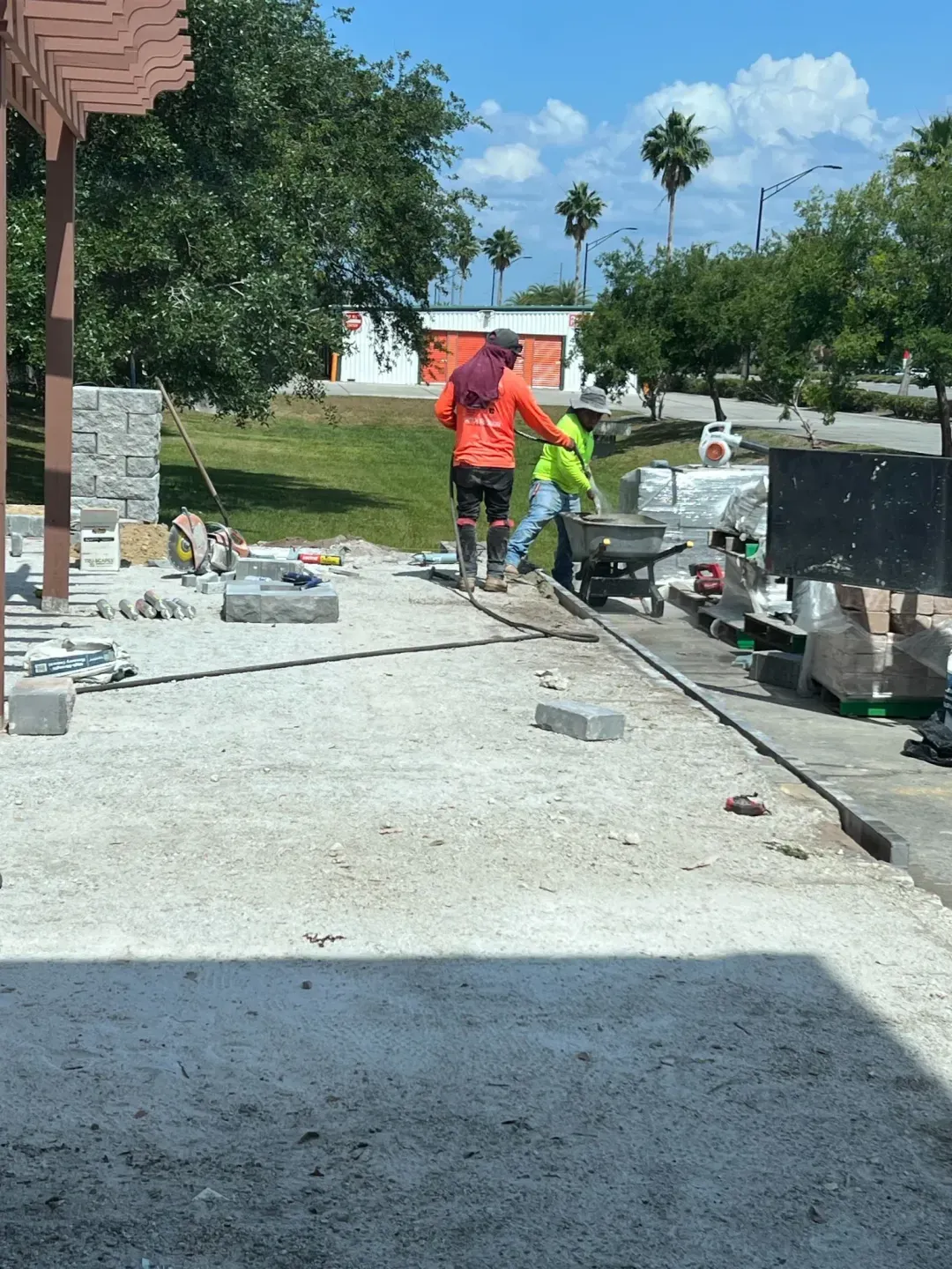 Crew preparing the base for a commercial paver installation, wheelbarrow and tools staged on site.