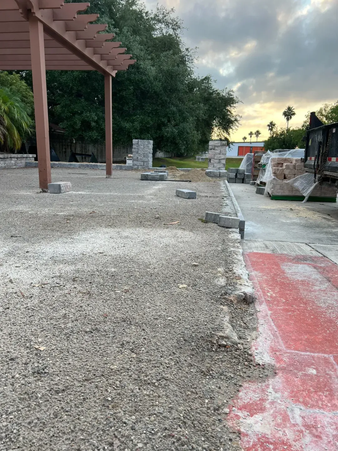 Evening job site with a cedar pergola frame in place and the paver base being prepped beneath it.