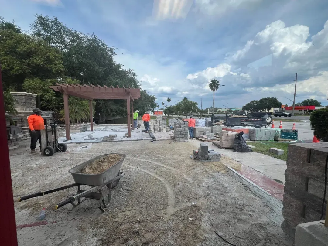 Crew in safety orange building a commercial paver plaza and pergola, with materials staged around the site.