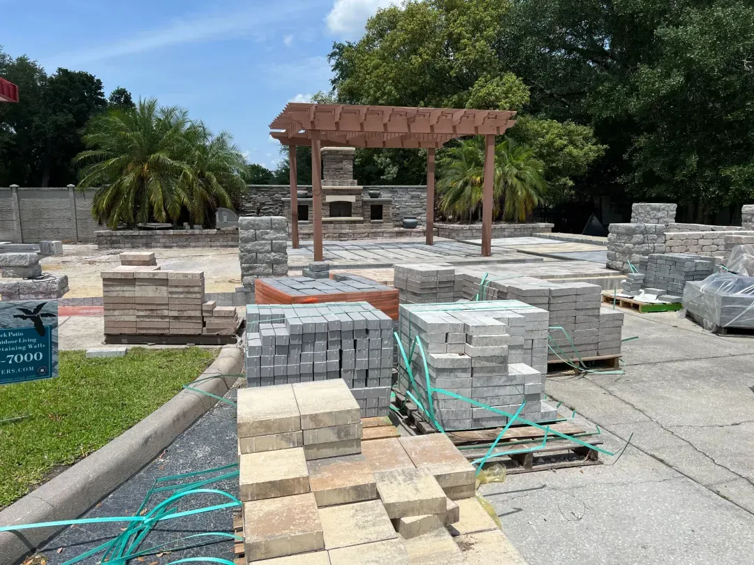 Pallets of pavers staged in front of a finished pergola and outdoor fireplace on a commercial job site.