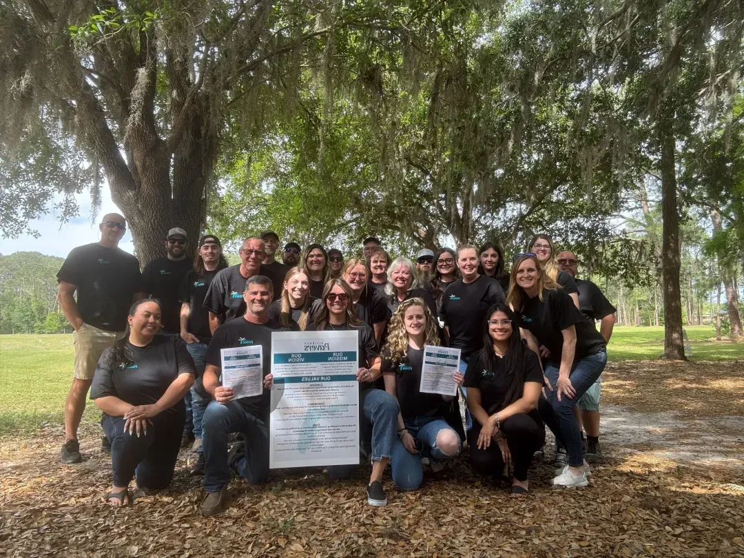 The full Alliance Pavers team gathered outdoors, holding a printed copy of the company's vision, mission, and values.